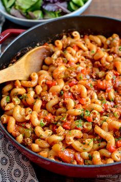 a skillet filled with pasta and vegetables on top of a wooden table next to a salad