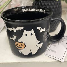 a black coffee mug with a ghost and pumpkin on it sitting next to a potted plant