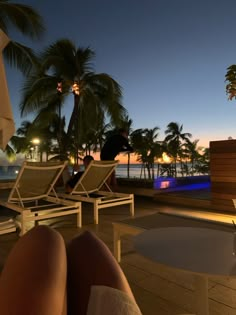 a person sitting on a deck next to some chairs and palm trees at night with the ocean in the background