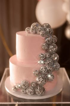 a three tiered pink cake with silver balls on it's side and balloons in the background