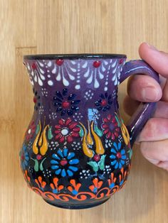 a hand holding a colorful painted vase on top of a wooden table next to a purple cup