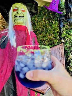 a person holding a drink in front of a fake witch with white hair and green eyes