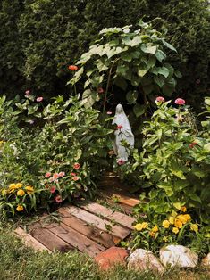 a statue in the middle of a garden surrounded by flowers and plants, with an arch made out of wood planks