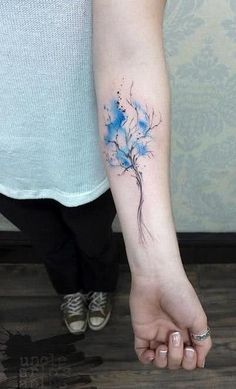 a blue flower tattoo on the left inner arm and wrist is shown in front of a woman's right hand