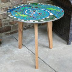 a mosaic table sitting on top of a cement floor next to a chair and brick wall