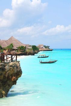 two boats floating in the ocean next to a rocky cliff with thatched huts on it