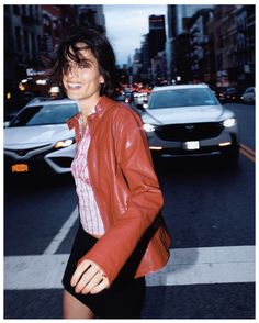 a woman is crossing the street in front of some cars and buildings at night, wearing a red leather jacket