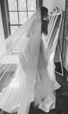 a woman in a wedding dress standing next to a window and holding a veil over her head