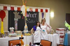 two women in aprons standing behind a table with cakes and desserts on it