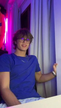 a young man wearing glasses sitting in front of a purple light with his hand on the wall