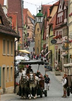 two horses pulling a cart down a narrow street with people walking on the sidewalk and buildings in the background