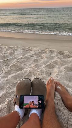 a person sitting in the sand with their feet up and holding a cell phone to take a photo