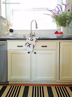 a kitchen with white cabinets, black and white striped area rug and flowers in the window