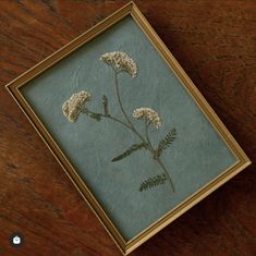 a painting with white flowers in a gold frame on a wooden table next to a cell phone