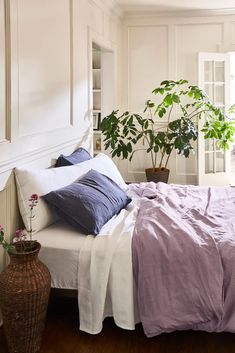 a bed with purple and white linens in a bedroom next to a potted plant