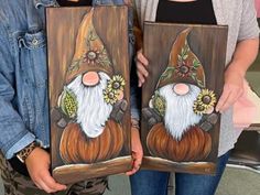 two women are holding paintings with gnomes painted on them, one has sunflowers in her hair and the other is wearing a denim jacket