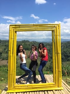 three women standing in front of a yellow frame with the sky and trees behind them