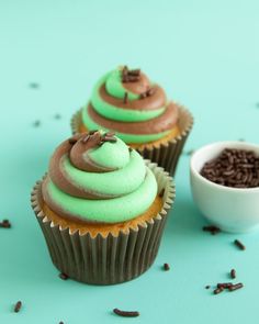 two cupcakes with green frosting and chocolate sprinkles on a blue surface