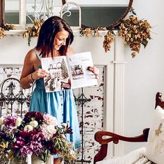 a woman standing in front of a mirror reading a magazine with flowers on the table