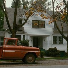 an old red truck parked in front of a white house with trees and bushes around it