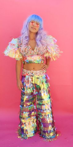 a woman with blue hair and glasses in a colorful outfit standing against a pink background