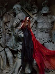 a woman in a red and black dress standing next to some statues with armor behind her