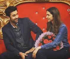 a man and woman sitting on top of a red couch next to each other holding flowers