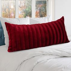 a red pillow sitting on top of a white bed next to two windows with trees in the background