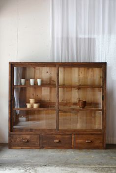 a wooden cabinet with glass doors and drawers in front of a white curtained window