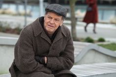 an older man sitting on a park bench wearing a hat and coat with his arms crossed