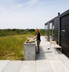 a person standing at a table with food in front of a black building and grassy field