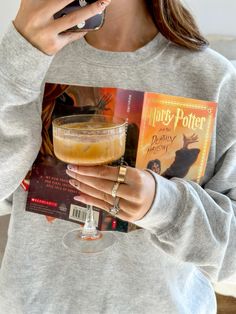 a woman holding up a harry potter book and a drink in her hand while looking at her phone