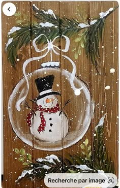 a snowman in a glass ornament hanging from a wooden fence with pine branches