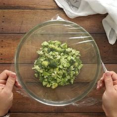 a person holding a glass bowl filled with chopped broccoli on top of a wooden table