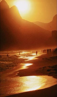 people are walking on the beach as the sun goes down in the mountains behind them