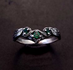 a silver ring with green stones and leaves on the sides, sitting on a black surface