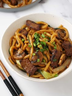 a white bowl filled with noodles and meat on top of a table next to chopsticks