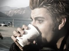 a man drinking from a white cup with mountains in the background and water behind him