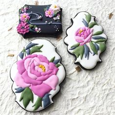 three decorated cookies sitting on top of a white table next to a black bag and pink flowers