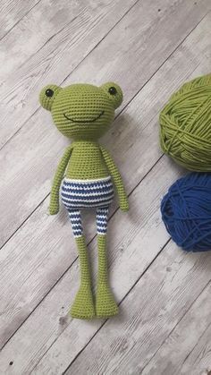 two balls of yarn next to a green frog stuffed animal on a wooden floor with white planks