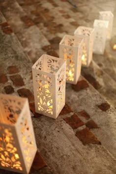 four white lanterns sitting on top of a stone floor next to each other with lit candles in them