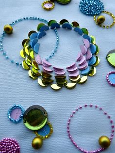 many different colored beads and necklaces on a white tablecloth with blue, pink, green, yellow, and purple colors