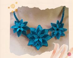 a blue crocheted necklace with flowers on it and a piece of paper behind it