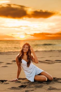 a beautiful young woman sitting on top of a sandy beach next to the ocean at sunset