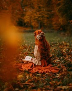 a woman sitting on top of a field covered in leaves reading a book next to trees