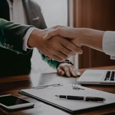 two people shaking hands over a desk with a laptop and notebook on it, while another person holds the hand of another person's hand