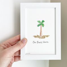 a hand holding up a small white frame with a green palm tree on it that says our beach house