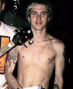 a shirtless man holding a guitar in his right hand and another person with tattoos on their left arm