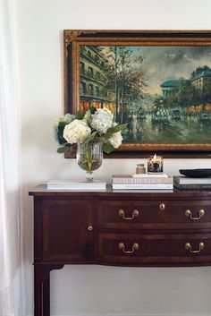 a painting hangs on the wall above a dresser with flowers and books in front of it