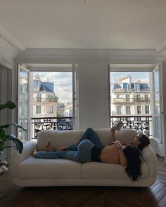 a woman laying on top of a couch in front of two large windows next to a potted plant